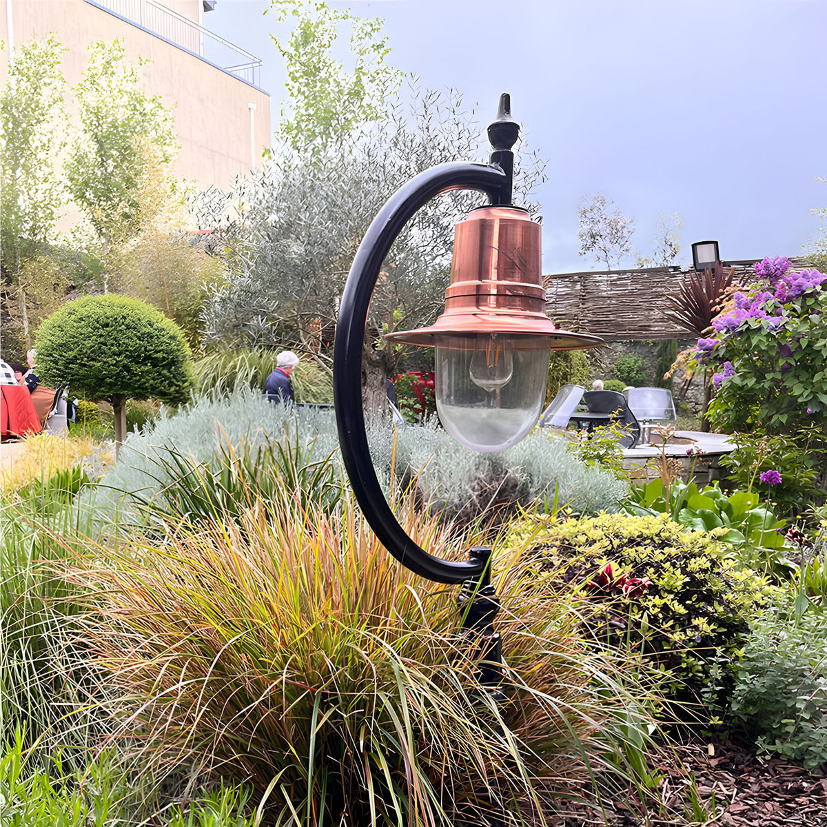 Large Vintage Cast Iron Pedestal Post Light with copper teardrop shade and curved black gooseneck arm, shown installed in a garden bed among ornamental grasses.