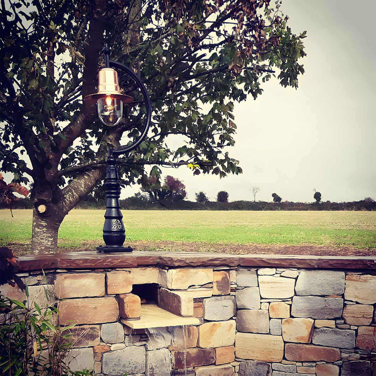 Large vintage cast iron pedestal post light with copper teardrop shade and gooseneck arm, mounted on a stone wall beside a tree, overlooking a rural field.