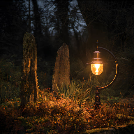 Large Vintage Cast Iron Pedestal Post Light in black with curved goose-neck arm and teardrop glass shade, glowing beside stone markers in an outdoor garden setting at dusk.