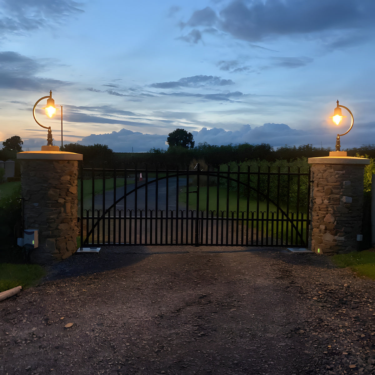 Large Vintage Cast Iron Pillar Light in black, gooseneck curve with teardrop shade, illuminated atop stone gate piers at dusk.