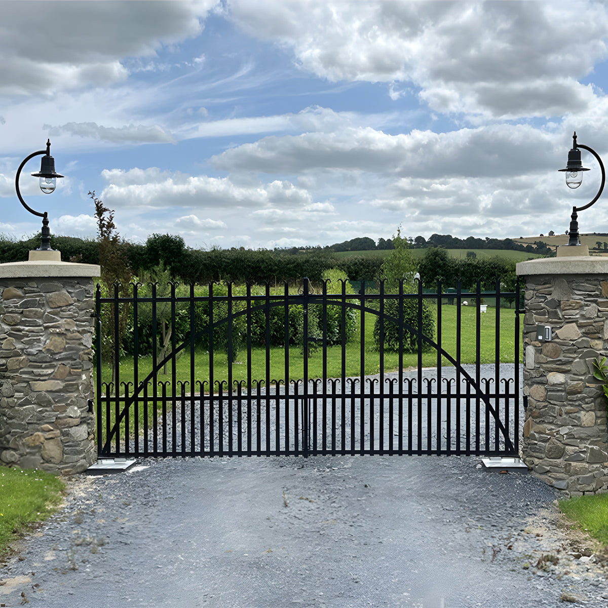 Large vintage cast iron pillar lights with curved goose neck arms and teardrop shades mounted on stone gate piers, flanking a black metal driveway gate.