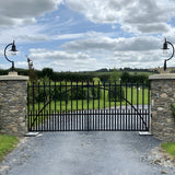 Large vintage cast iron pillar lights with curved goose neck arms and teardrop shades mounted on stone gate piers, flanking a black metal driveway gate.