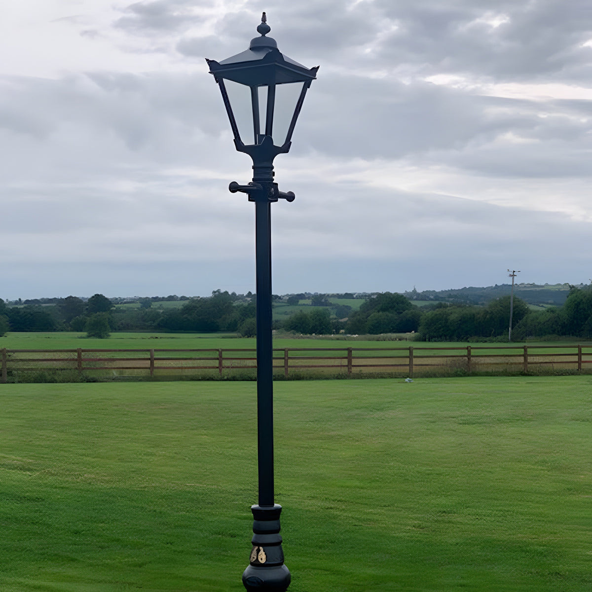 Victorian Manor 3m cast iron lamp post in black with hexagonal lantern head and glass panels, shown outdoors on a lawn beside a field.