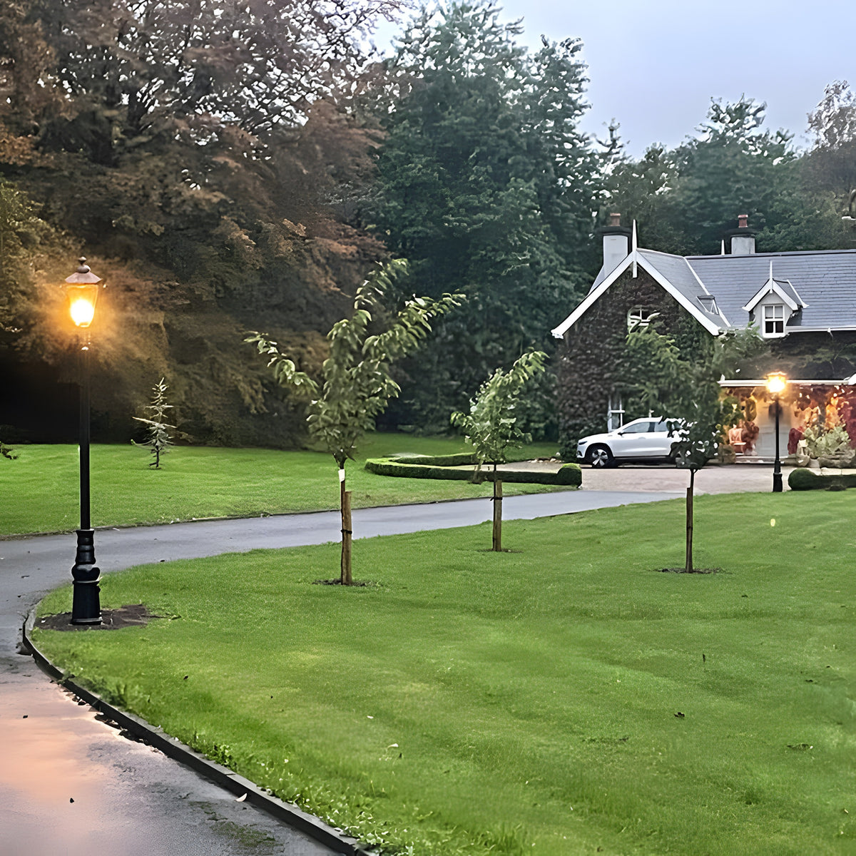 Victorian Manor cast iron lamp post in black, 3m traditional design with lantern-style glass shade, shown illuminated on a garden driveway edge at dusk.
