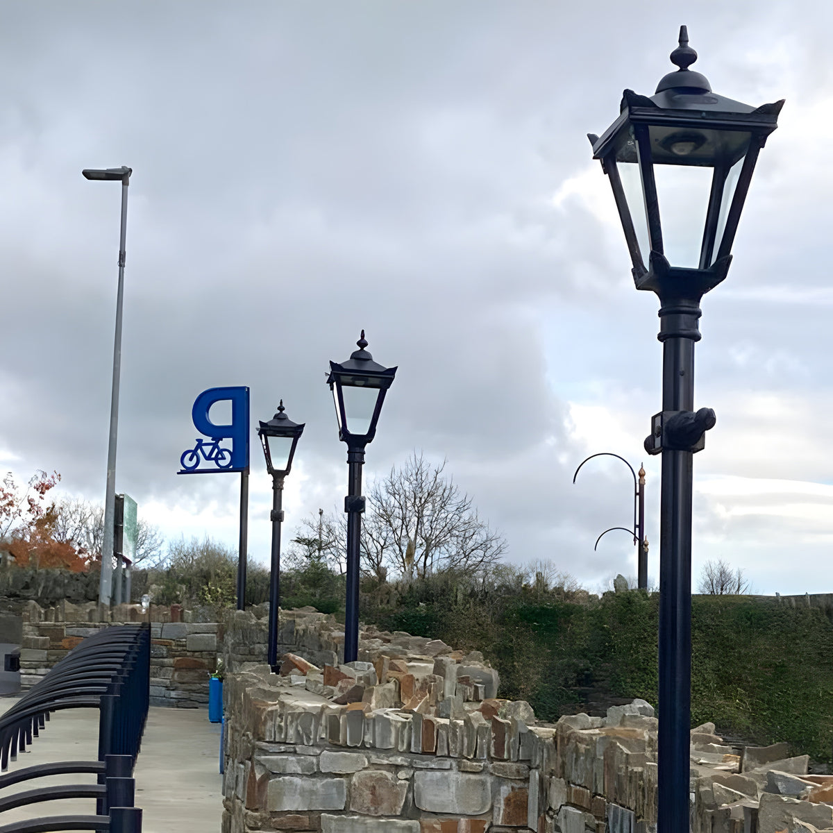 Victorian Manor Cast Iron Lamp Post in black, shown outdoors along a stone wall pathway with matching lamp posts and a bicycle parking sign in the background.