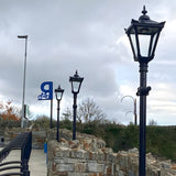 Victorian Manor Cast Iron Lamp Post in black, shown outdoors along a stone wall pathway with matching lamp posts and a bicycle parking sign in the background.