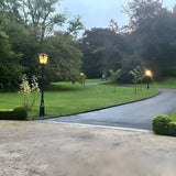 Victorian Manor 3m cast iron lamp post in black with lantern-style glass head, illuminated on a garden driveway lined with lawn and trees.