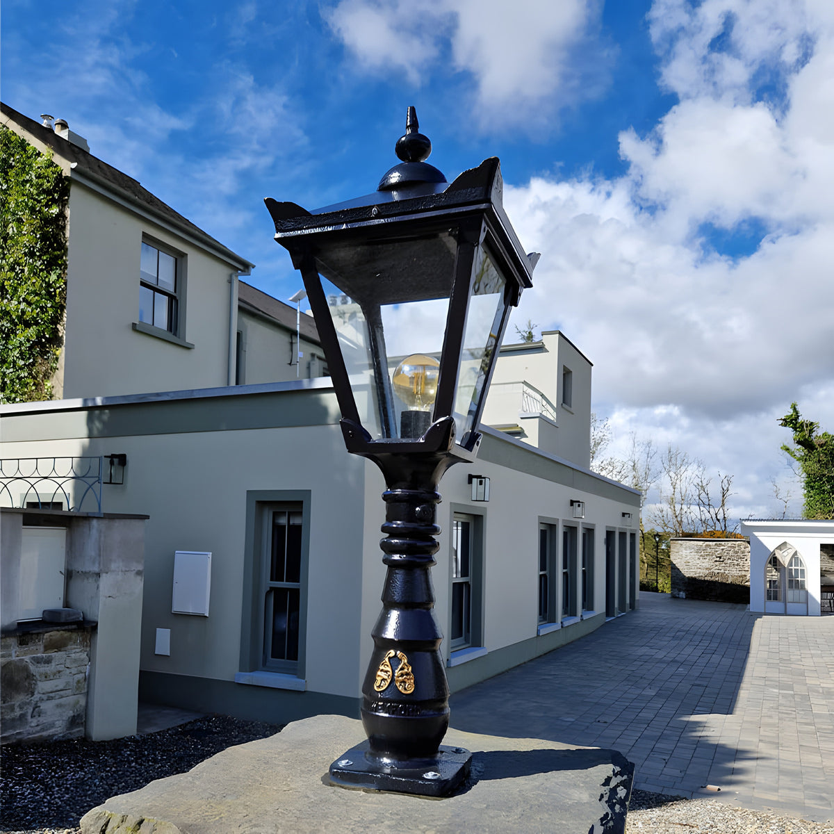 Small Victorian Manor cast iron pedestal post light in black, with square lantern head, glass panels, exposed globe bulb, and ribbed column, mounted outdoors on a stone base by a building.