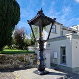 Small Victorian Manor cast iron pedestal post light in black with square glass lantern, mounted on stone base outside a house exterior.