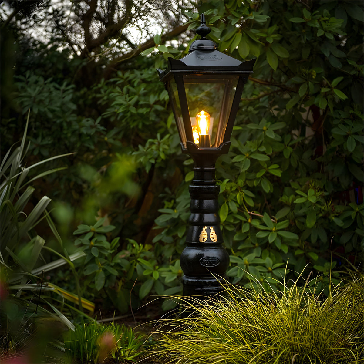 Large Victorian Manor cast iron pedestal post light in black, slim column with lantern head and glass panels, glowing bulb, set in a garden among greenery.