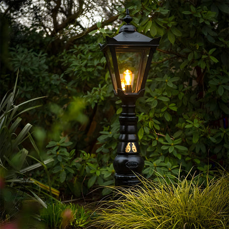 Large Victorian Manor cast iron pedestal post light in black, slim column with lantern head and glass panels, glowing bulb, set in a garden among greenery.