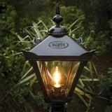 Large Victorian Manor cast iron pedestal post light in black, traditional lantern-style head with glass panels and lit E27 bulb, set against garden greenery.