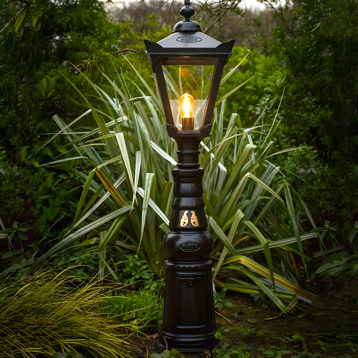 Large Victorian Manor cast iron pedestal post light in black, glass lantern top with glowing bulb, slim column base, shown outdoors amid garden foliage.