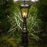 Large Victorian Manor cast iron pedestal post light in black, glass lantern top with glowing bulb, slim column base, shown outdoors amid garden foliage.