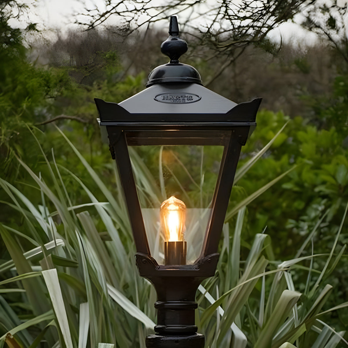 Large Victorian Manor cast iron pedestal post light in black, traditional lantern-style design with glass panels and lit E27 bulb, shown outdoors amid garden foliage.