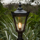 Large Victorian Manor cast iron pedestal post light in black, traditional lantern-style design with glass panels and lit E27 bulb, shown outdoors amid garden foliage.