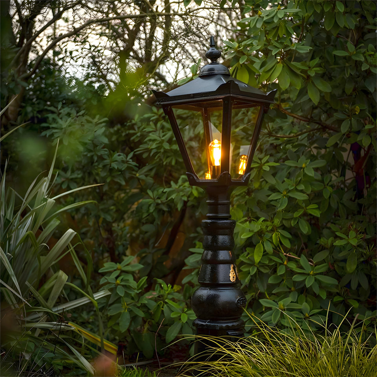 Large Victorian Manor cast iron pedestal post light in black, lantern-style head with glass panels and lit bulb, slim column base, set outdoors amid garden foliage.