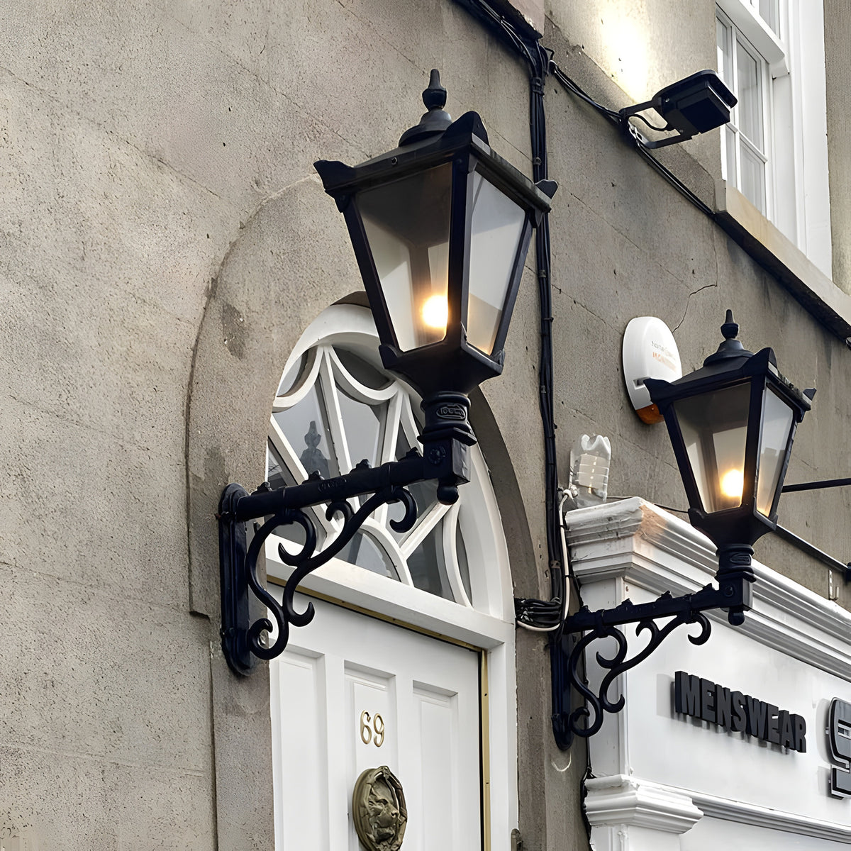 Victorian Manor wall lantern in black on a scroll bracket, lit and mounted beside an arched doorway on a stone building exterior.