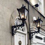 Victorian Manor wall lantern in black on a scroll bracket, lit and mounted beside an arched doorway on a stone building exterior.
