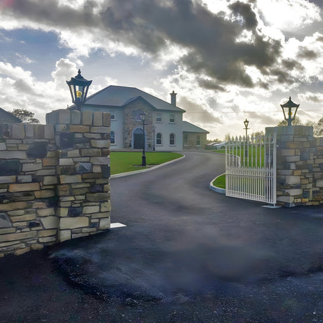 Black cast iron Victorian Manor pillar light mounted on stone gate piers, traditional lantern design with glass panels, framing a driveway to a country house.