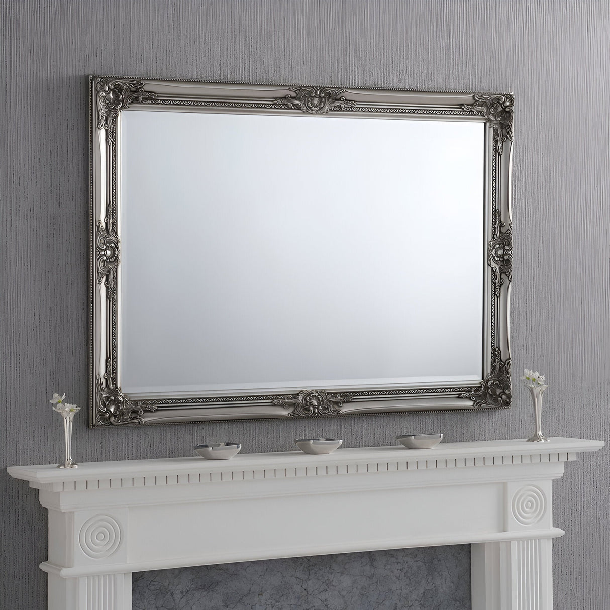 The Orbit Rectangle Wall Mirror Silver - 104x74cm, with its versatile design and ornate silver frame, is mounted on a textured gray wall. Beneath it, a white mantelpiece embellished with decorative molding displays three small vases filled with white flowers.