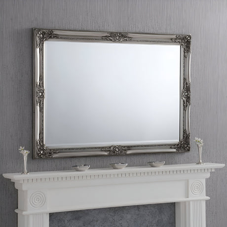 The Orbit Rectangle Wall Mirror Silver - 104x74cm, with its versatile design and ornate silver frame, is mounted on a textured gray wall. Beneath it, a white mantelpiece embellished with decorative molding displays three small vases filled with white flowers.