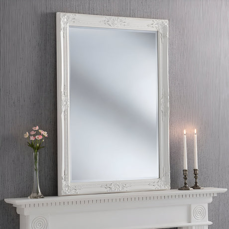 An expansive and ornate Orbit Rectangle Wall Mirror White - 104x74cm graces the space above a white fireplace mantel. Atop the mantel rest a vase filled with pink flowers and two lit candlesticks. The backdrop is a softly textured grey wall, perfectly combining elegance with contemporary style.