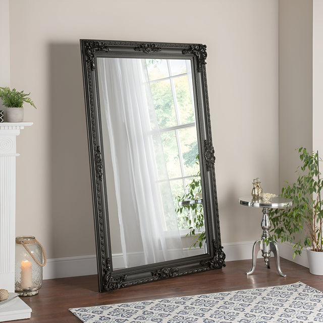 A Hilton Leaner Mirror Black - 178x117cm with a decorative black frame is propped against a light-colored wall in a cozy room. A window with sheer curtains is reflected in the mirror. To the right, there's a small round table with a plant resting on a wooden floor covered by a patterned rug.