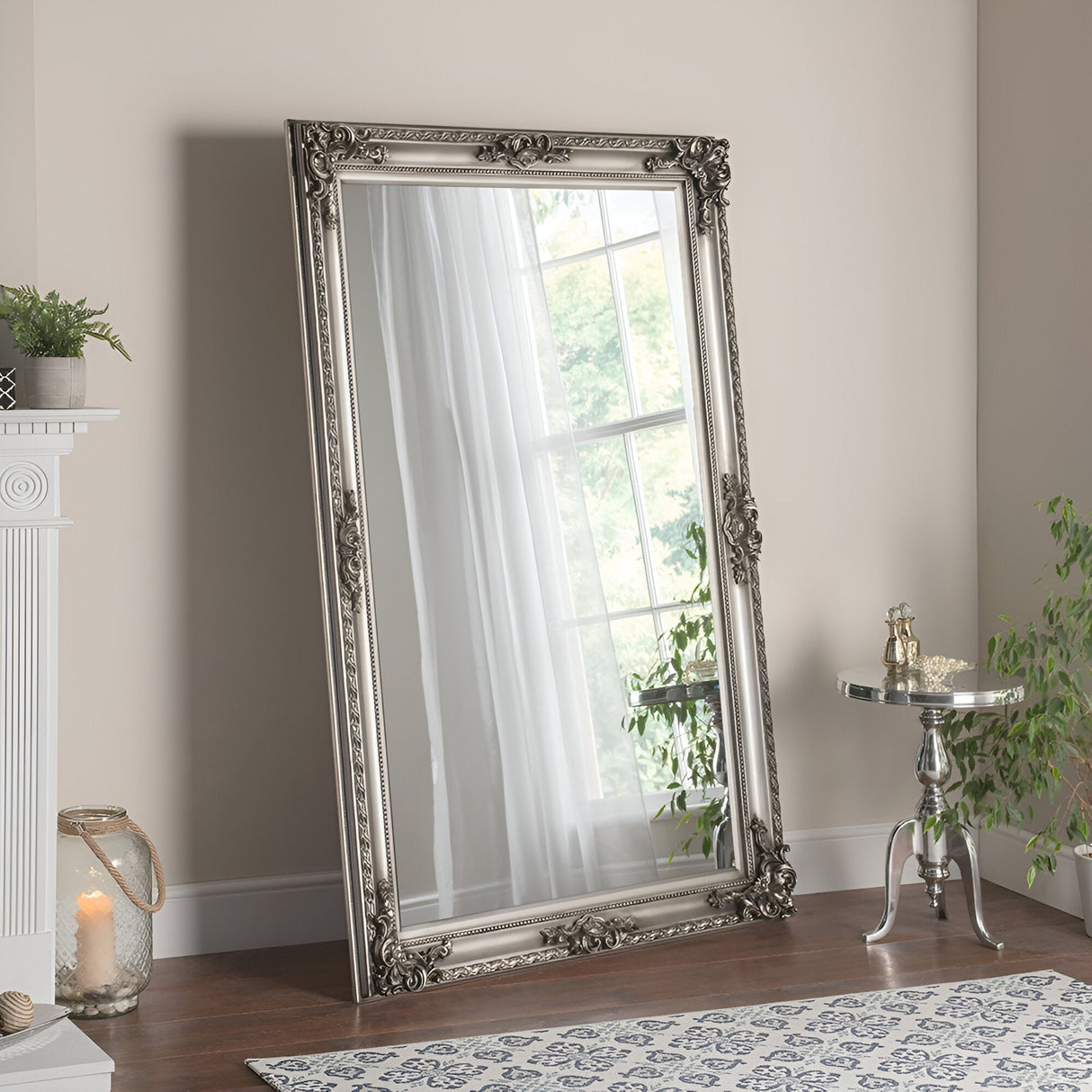 The Hilton Leaner Mirror Silver, measuring 178x117cm, leans against a beige wall, mirroring a window dressed in sheer curtains. Nearby, a small silver table adorned with decorative items stands next to a plant. A softly glowing lit candle on the floor beside the fireplace adds to the minimalist design.