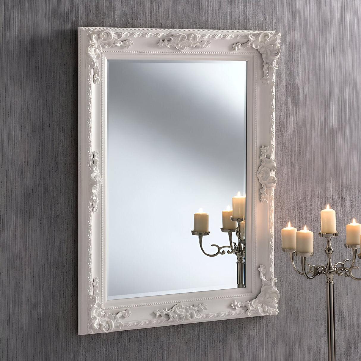 The Hilton Rectangle Wall Mirror in White (120x90cm) adorns a textured grey wall with its ornate frame. Gentle lighting is reflected in the mirror, while the glow of a candelabra with lit candles complements the timeless design, infusing elegance into this exquisite home decor piece.