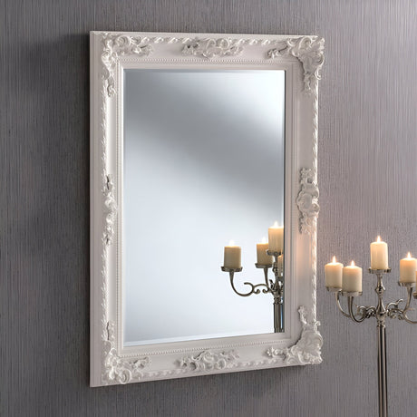 The Hilton Rectangle Wall Mirror in White (120x90cm) adorns a textured grey wall with its ornate frame. Gentle lighting is reflected in the mirror, while the glow of a candelabra with lit candles complements the timeless design, infusing elegance into this exquisite home decor piece.
