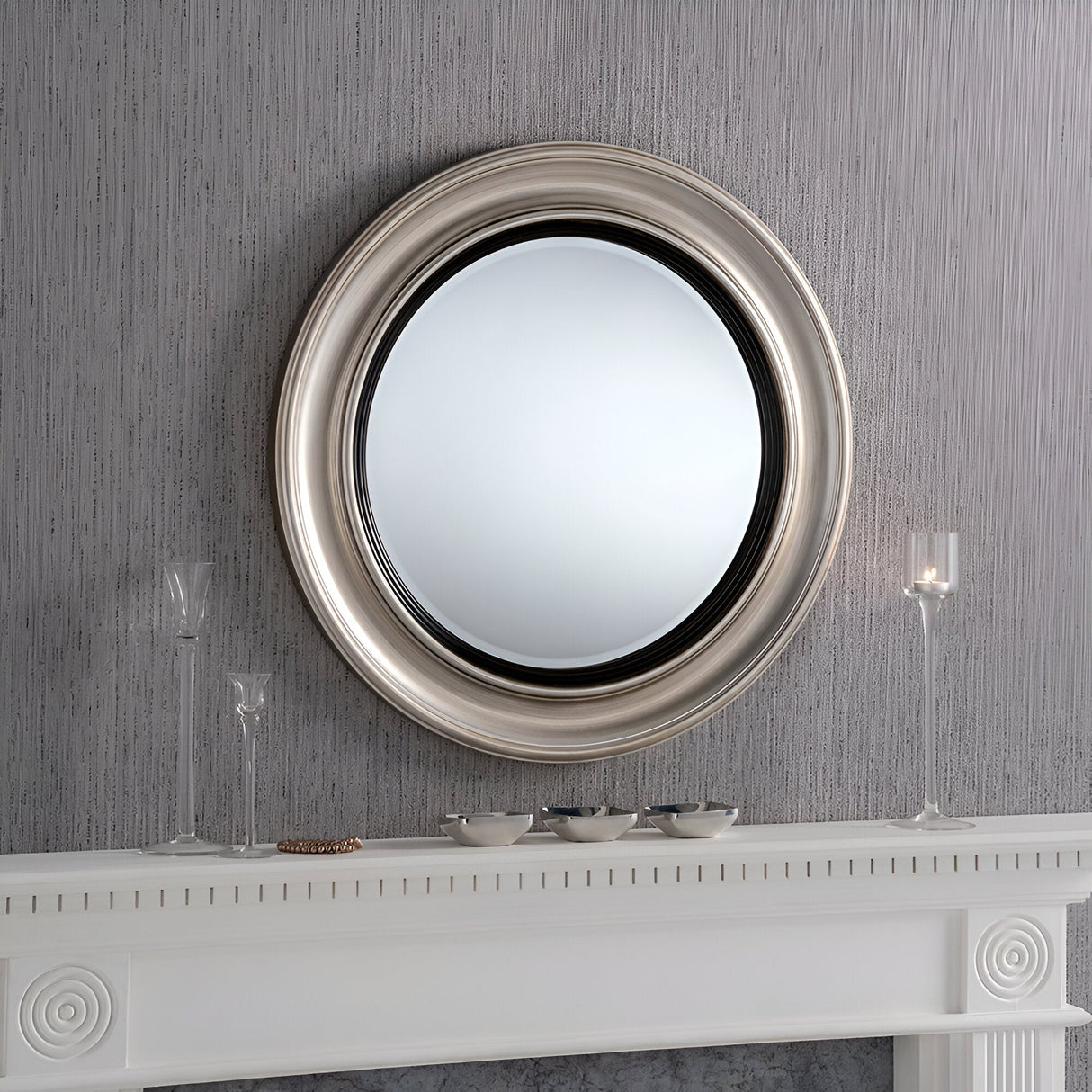 The Hamilton Circle Wall Mirror in Silver/Black with a 66cm diameter brings a contemporary flair as it hangs above the white mantel. The mantel's modern elegance is highlighted by three glass candlesticks and several small bowls set against the textured gray wall.