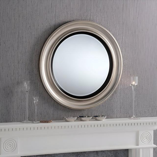 The Hamilton Circle Wall Mirror in Silver/Black with a 66cm diameter brings a contemporary flair as it hangs above the white mantel. The mantel's modern elegance is highlighted by three glass candlesticks and several small bowls set against the textured gray wall.