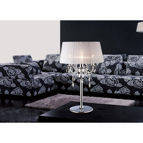 Olivia 3 Light E14 Table Lamp in polished chrome with crystal drops and a white pleated shade, shown on a coffee table beside a black and white floral sofa.