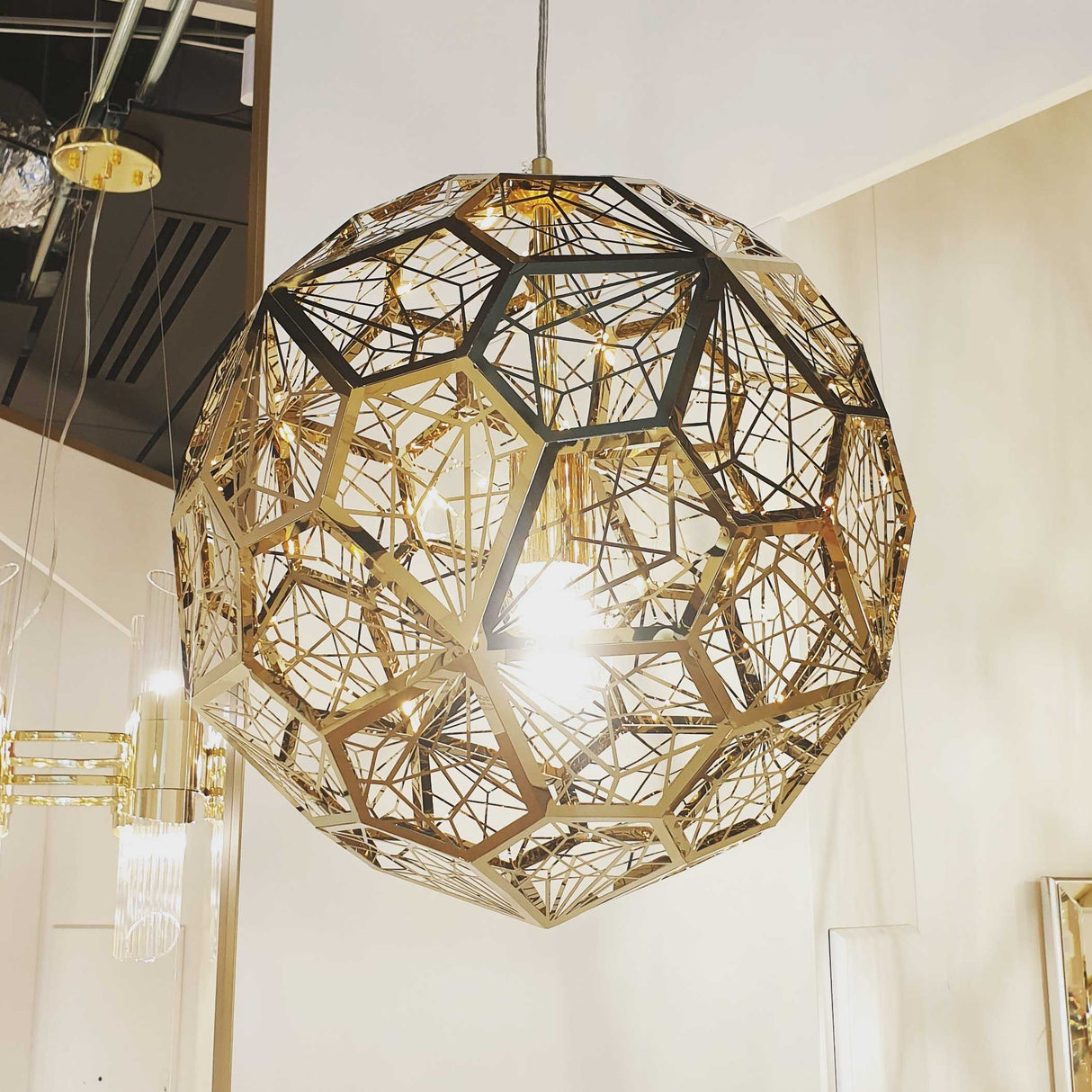 The Rango Acid Small Pendant Light in Brass embodies a contemporary style with its spherical design made of intricate, overlapping golden metal polygons. It hangs elegantly from the ceiling, providing ambient lighting that shines brightly from the center and casts patterned shadows on nearby surfaces.