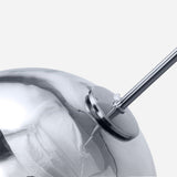 A close-up view showcasing the metallic, mirror-like Grand Ball Glass Pendant in silver, measuring 25 cm, with a precision instrument attached to its side. The scene is enhanced by LED compatible lighting against a plain light gray background, highlighting its modern aesthetic.