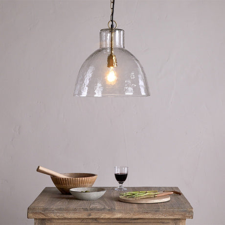 Inayah Recycled Glass Pendant Light in Clear, a hammered dome shade with brass fittings and black chain, shown above a wooden table set with bowls and a wine glass.