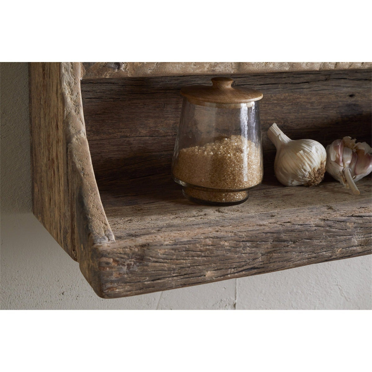 Ibo Reclaimed Wood Wall Shelf in natural finish, close-up of lower tier with raised front edge holding a lidded glass jar of sugar and garlic bulbs.