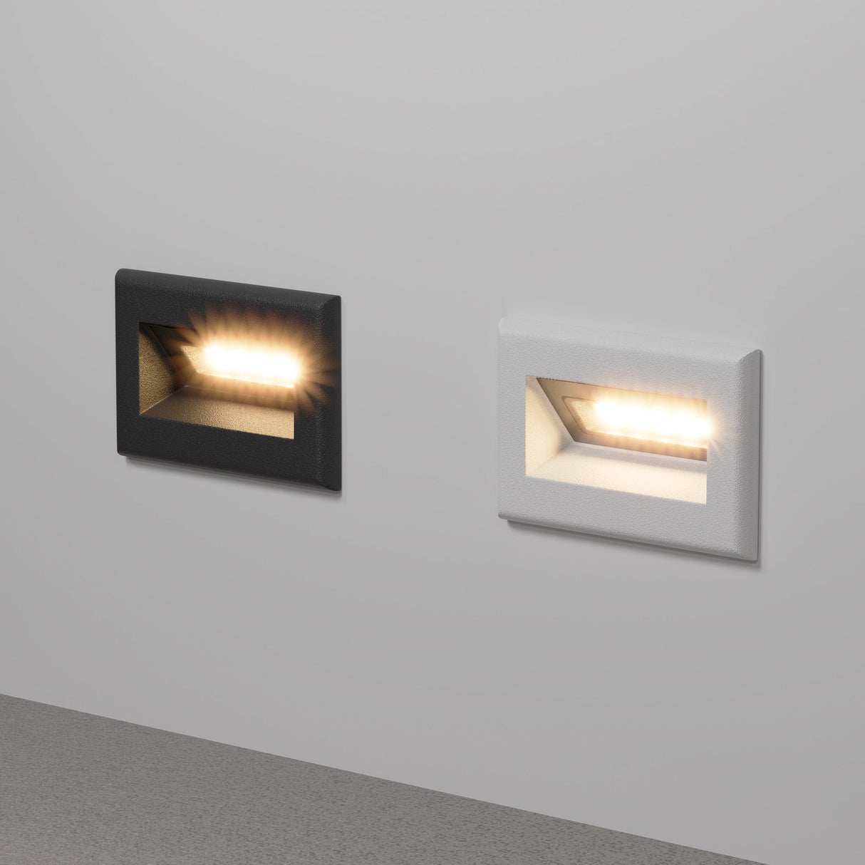 Two Bosca Outdoor LED Recessed Wall Lights in black frames are mounted on a light gray wall, emitting a warm glow. Their energy-efficient LEDs illuminate the area in front, casting light on the floor below and highlighting their sleek, modern design.