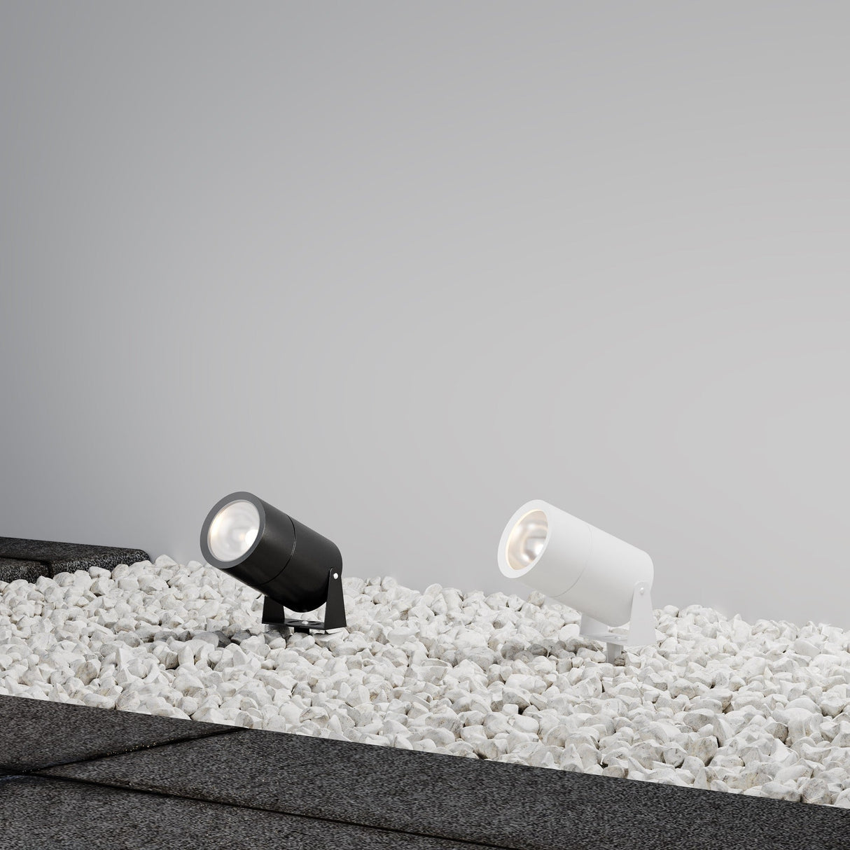 Two Bern Outdoor LED Spike Lights, 15W 3000K in graphite, are placed on white pebbles against a grey wall. Angled upward, they cast a soft glow with dark paving stones framing the pebble area.