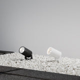 Two Bern Outdoor LED Spike Lights, 15W 3000K in graphite, are placed on white pebbles against a grey wall. Angled upward, they cast a soft glow with dark paving stones framing the pebble area.