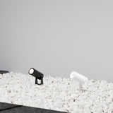 Two small, cylindrical Bern Outdoor LED Spike Lights (5W, 3000K) in graphite are energy-efficient LEDs mounted among white pebbles against a plain grayish background.