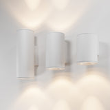The Shim Outdoor Wall Light 24W 3000K - White enhances spaces with three cylindrical wall-mounted fixtures offering minimalist design. They provide soft, warm LED light upwards and downwards. IP65 rated for outdoor use, these create a modern, symmetrical aesthetic against a white wall.