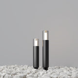 Two Glide Outdoor Long Spike Lights in graphite, featuring modern black design and clear tops, stand on white stones against a plain gray background. These lights softly illuminate the area, providing elegant outdoor lighting with their long spike design.