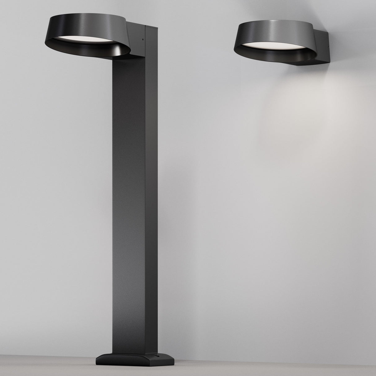 A Share LED Bollard Light 65cm in graphite stands beside a matching wall fixture, both with round shades illuminating a plain gray wall. Perfect for minimalist and contemporary settings, these fixtures beautifully complement outdoor spaces akin to sophisticated LED pathway lighting.