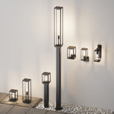 A minimalist and elegant outdoor setup features tall and small rectangular lantern-style lights, including the Cell Pedestal Light 22cm in Graphite, on pebble-covered ground. Wall-mounted lights on a white wall cast warm glows. This design has an IP54 rating for enhanced durability.