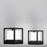 Two modern, black square lamps with white glowing interiors rest against a plain gray background. The Trim Pedestal Light 210mm in sleek graphite and aluminium is on the left, slightly taller than the right lamp. Ideal for stylish outdoor lighting solutions.