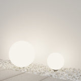 An Erda Pedestal Light 49cm - White emits a soft glow, nestled on white pebbles next to a tiled surface. This modern design complements the plain, light-colored wall, making it ideal for outdoor lighting.