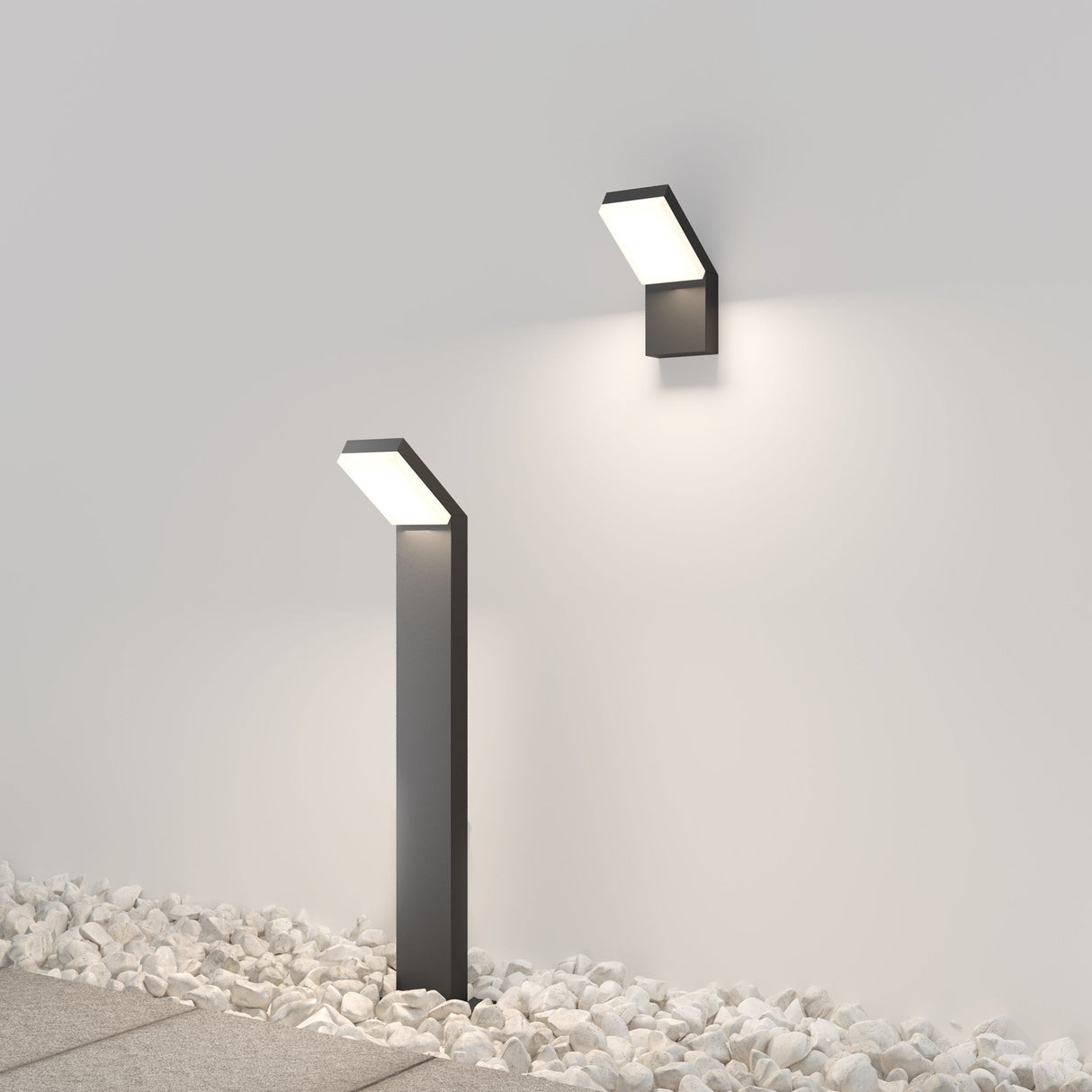 The Paso Outdoor LED Wall Light 3000K in black pairs with a similar freestanding design, illuminating white pebble-covered ground with a soft glow and casting light on the gray surface. Its chic rectangular shape is perfect for any stylish outdoor setting.