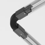 The 3-Phase Surface-Mounted Track Trinity Flexible Connector in black joins two silver articulated rails at a right angle with its ribbed, metallic design, perfect for modern interiors. The grooved rails are secured with black clips against a simple white backdrop.